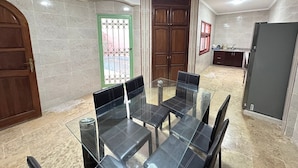A dining area with a glass table and chairs, a wooden door, a green-framed window, and a kitchen area with a refrigerator. - residence riad zitoune (Marrakech)