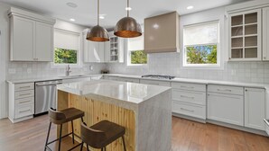 Een moderne keuken met witte kasten, een centraal eiland met krukken, hanglampen en grote ramen.