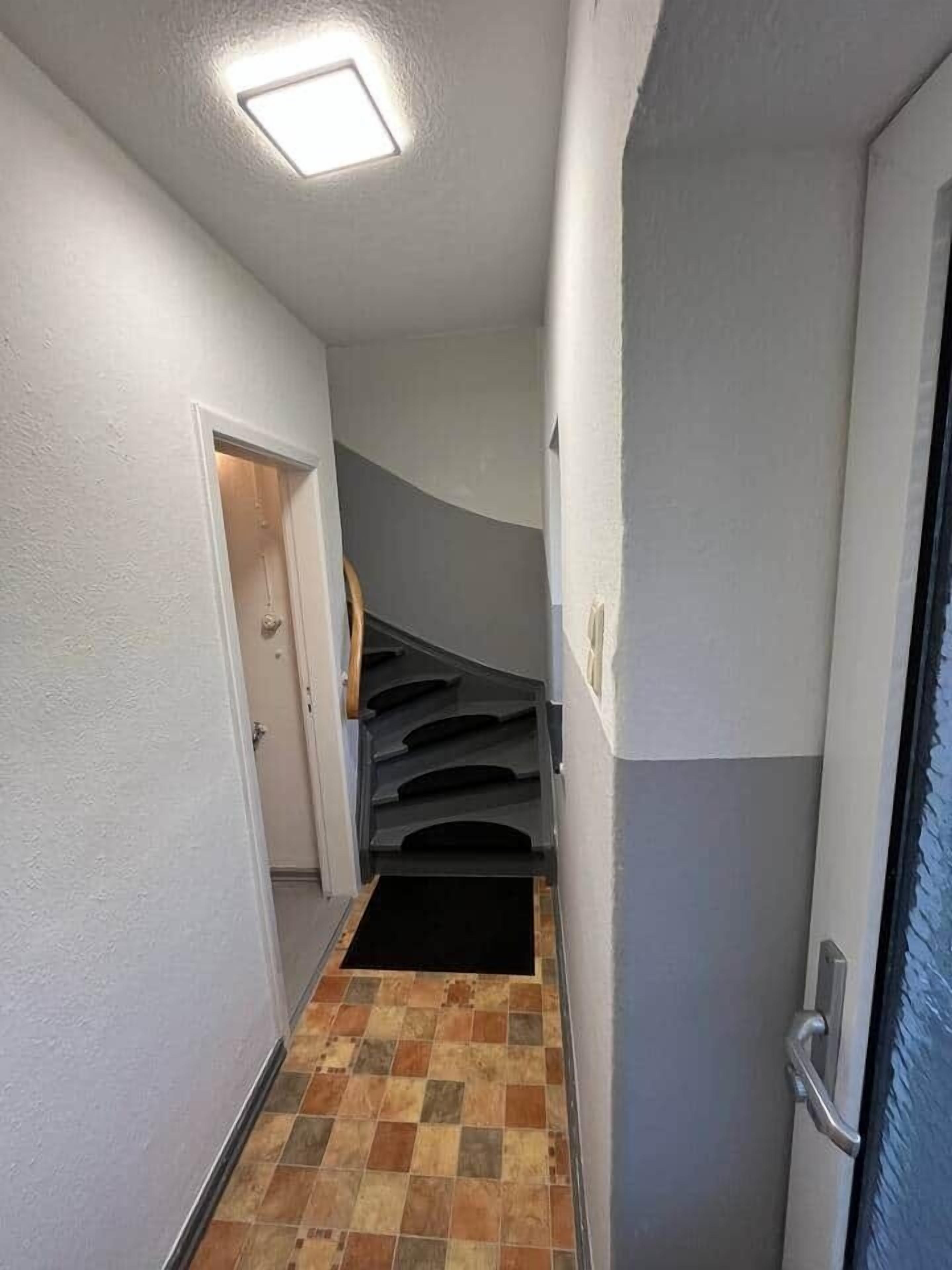 Un pasillo estrecho con suelo de baldosas, una puerta a la izquierda y una escalera a la derecha.