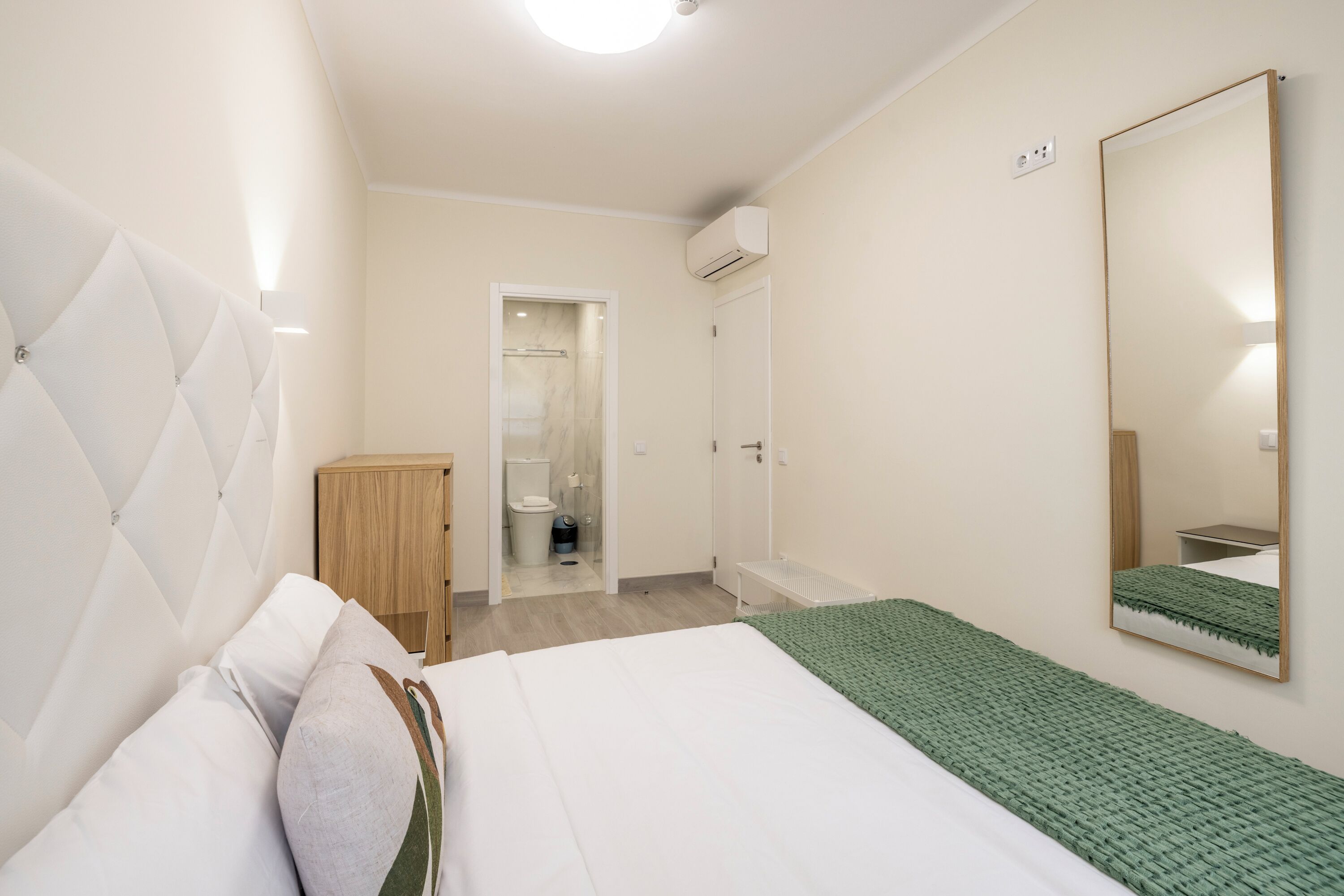 Une chambre à coucher avec un lit, un plaid vert, une armoire en bois et un miroir au mur.