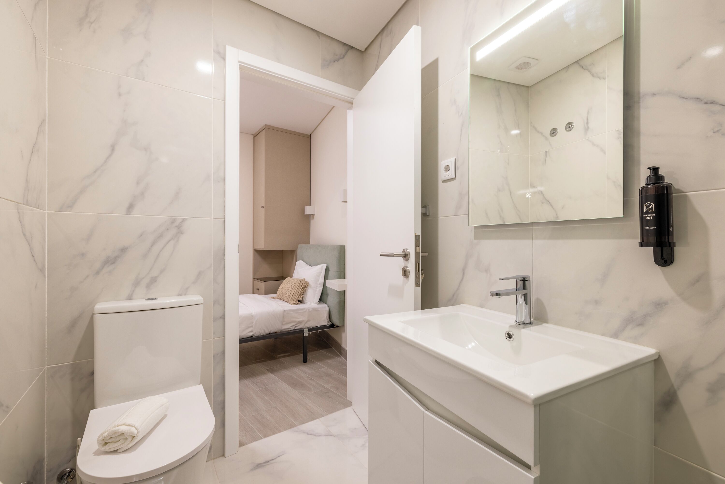 Un baño moderno con un lavabo blanco, un inodoro y una cama en la habitación contigua.