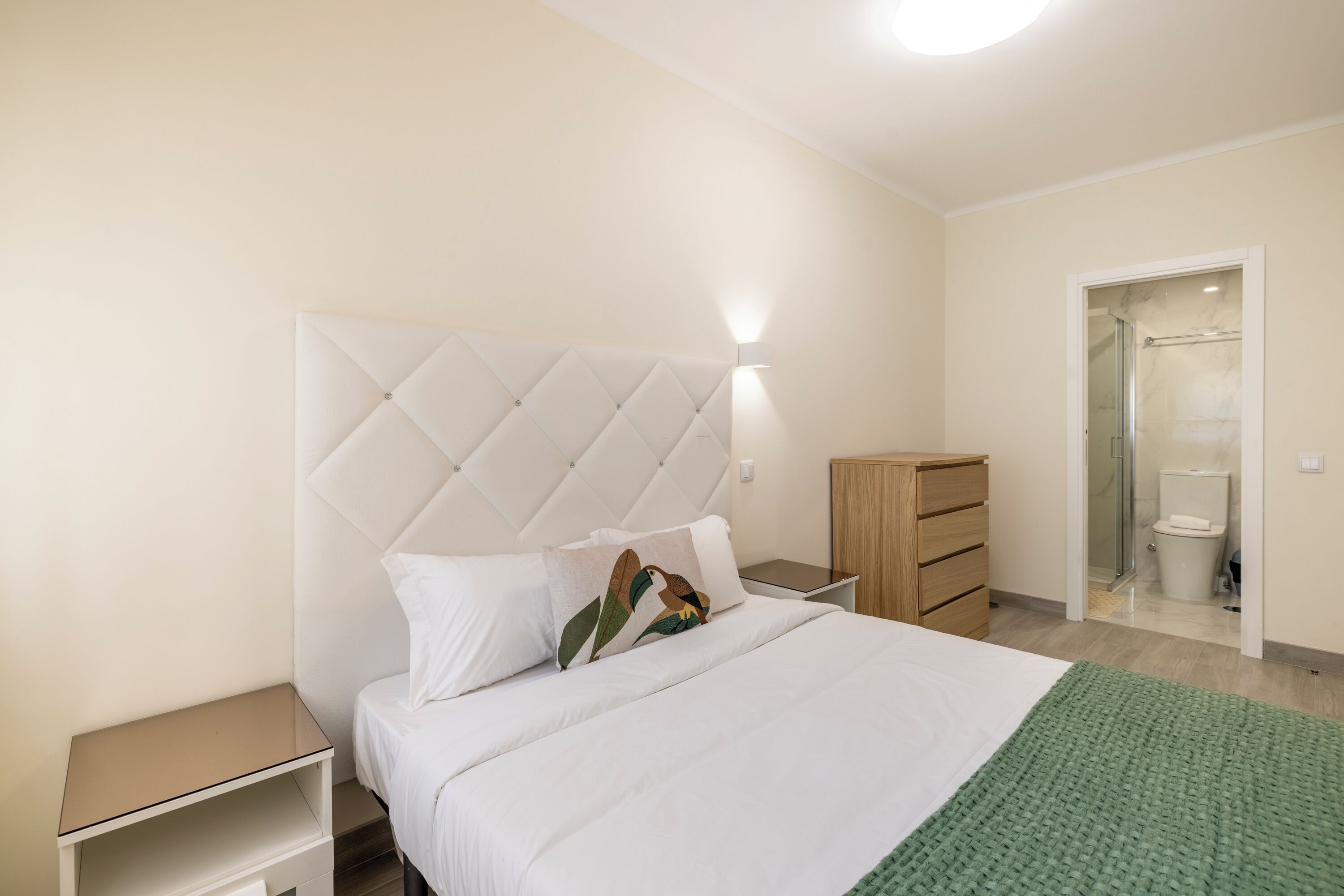 A bedroom with a tufted headboard, a bed with white bedding, a green throw, a wooden nightstand, a drawer unit, and a wooden wardrobe.