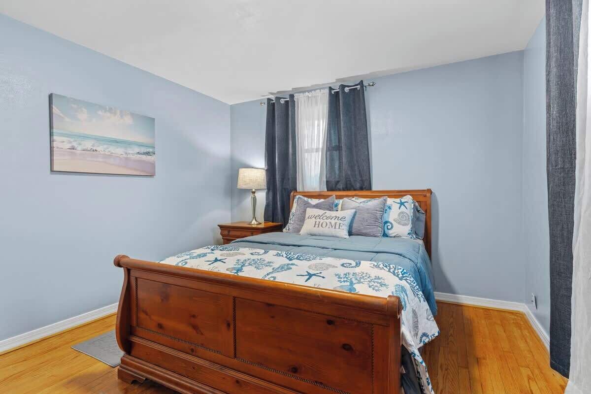 A bedroom with a wooden bed, a nightstand with a lamp, a beach-themed painting, and a window with curtains.