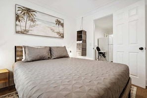A bedroom with a large bed, a painting of palm trees, a wardrobe, and a door leading to another room.