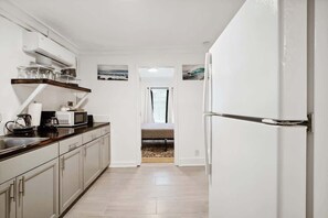 A modern kitchen with white cabinets, a wooden countertop, and a built-in microwave. There is a refrigerator on the right and a window with curtains in the background.
