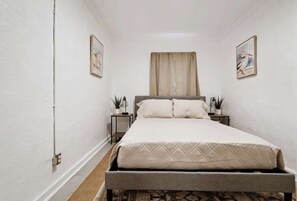 A narrow hotel room with a bed, two bedside tables, a window with curtains, and two wall-mounted paintings. - Cozy guest suite w/ kitchen & parking (Pensacola)