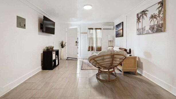 Une chambre d’hôtel moderne avec un plancher en bois, une chaise en rotin, un lit avec une tête de lit blanche, une table de chevet, un téléviseur fixé au mur et un tableau représentant des palmiers.