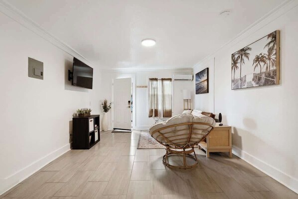 A modern hotel room with a wooden floor, a rattan chair, a bed with a white headboard, a bedside table, a TV mounted on the wall, and a painting of palm trees.