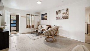 Un vaste espace de vie comprenant un canapé, un fauteuil en osier et une table basse. Une porte donne accès à un espace extérieur.