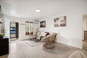 A spacious living area with a sofa, a wicker chair, and a coffee table. There is a door leading to an outdoor area.