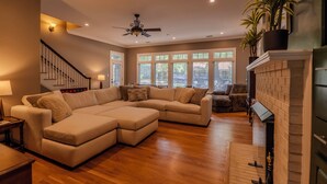 A spacious living room with a large sectional sofa, a fireplace, and a staircase with a ceiling fan. - 7BR/10BA Five Points Private Gathering Home | Sleeps 26, Theatre, Golf Sim, Elevator & Outdoor Areas (Raleigh)