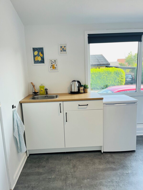 A modern kitchen with white cabinets, a wooden countertop, a kettle, a coffee maker, and a window with a view of a red car and greenery outside. - Cozy studio 500 m from the ferry and city center - perfect for 2 people (Hirtshals)
