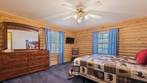 Une chambre à coucher de style chalet, avec une commode en bois, un lit recouvert d’une courtepointe, un ventilateur de plafond et un téléviseur fixé au mur.