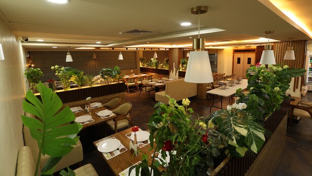A modern restaurant with wooden tables, white chairs, and green plants. - Hotel Pasupala Inn (Anantapur)