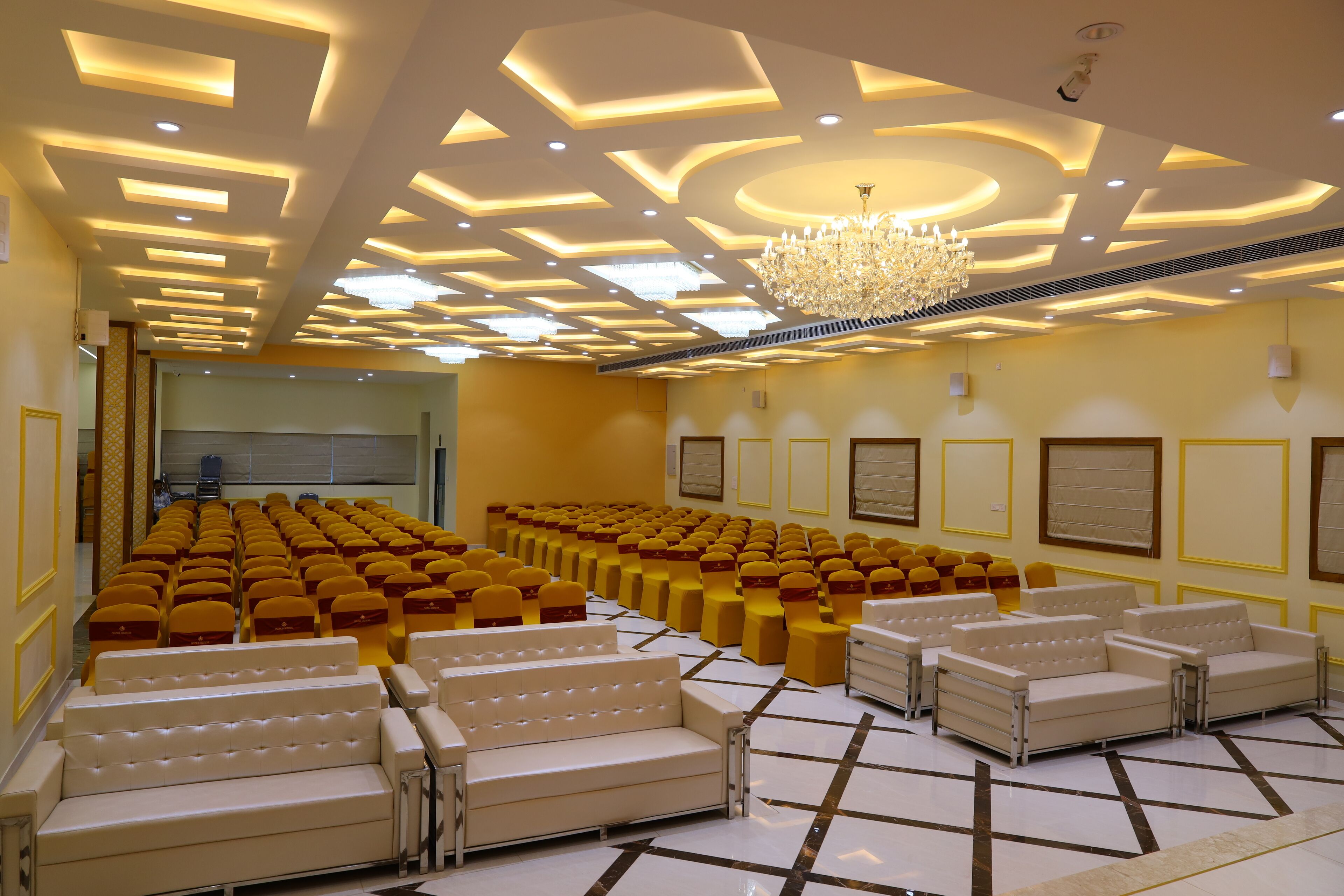Un salón de conferencias con hileras de sillas amarillas y sofás blancos, una araña y un techo estampado.