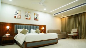 A hotel room with a large bed, a wooden headboard, two bedside tables with lamps, a sofa, a chair, and a ceiling fan. - Hotel Pasupala Inn (Anantapur)