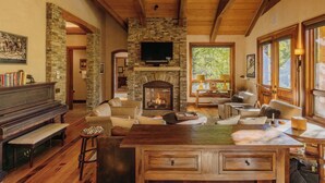 A cozy living room with a stone fireplace, wooden beams, a piano, and comfortable seating. - Cozy Cabin by InvitedHome | Theater Room, Bunk Room, Vaulted Ceilings (Mountain Village)