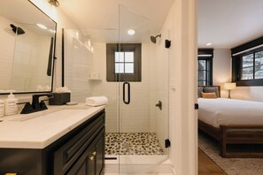 Ein modernes Badezimmer mit einer gläsernen Duschkabine, einem Waschtisch mit Toilettenartikeln und im angrenzenden Raum ein Bett mit einem Holz-Kopfbrett.