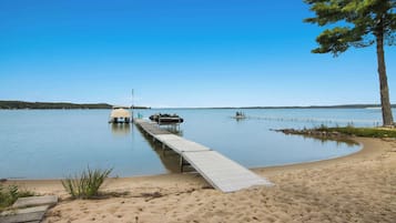 A sandy beach with a wooden dock extending into calm water, a picnic table, and a tree on the right.