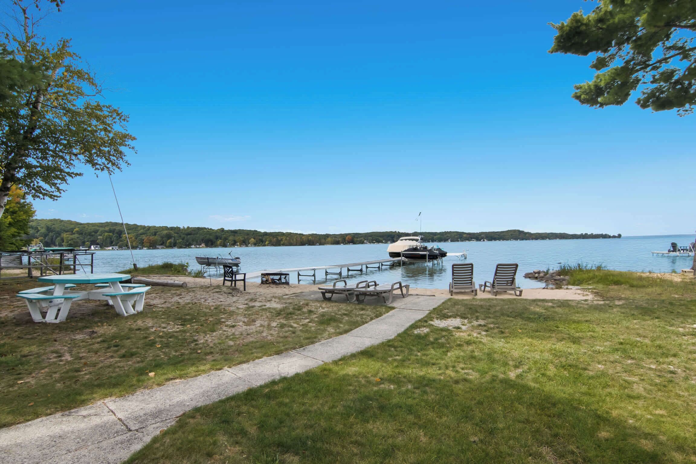 A lakeside picnic area with tables and chairs, a dock with a boat, and a clear view of the lake and distant shoreline.