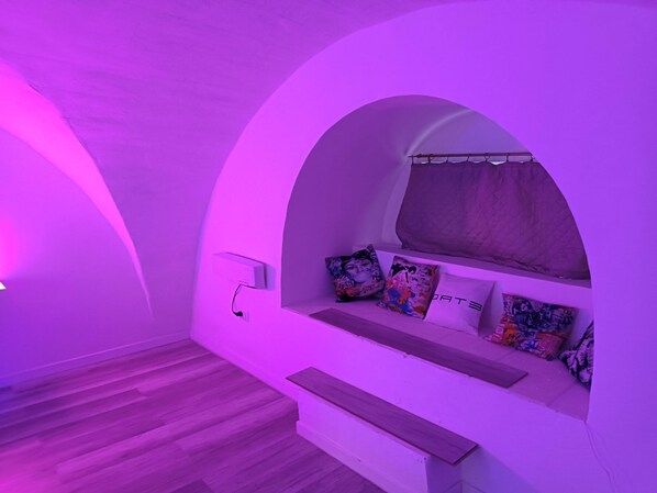 A room with a curved white wall, a bed with colorful pillows, a wooden bench, and a power outlet.