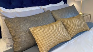 Een bed met een donkerblauwe hoofdbord, witte en grijze kussens, en een gouden kussen van gaas.