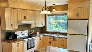 A modern kitchen with wooden cabinets, a central island with bar stools, a refrigerator, and a window with a view of trees. - Riverfront cottage-style home in the heart of the Acadian Peninsula (Pokemouche)