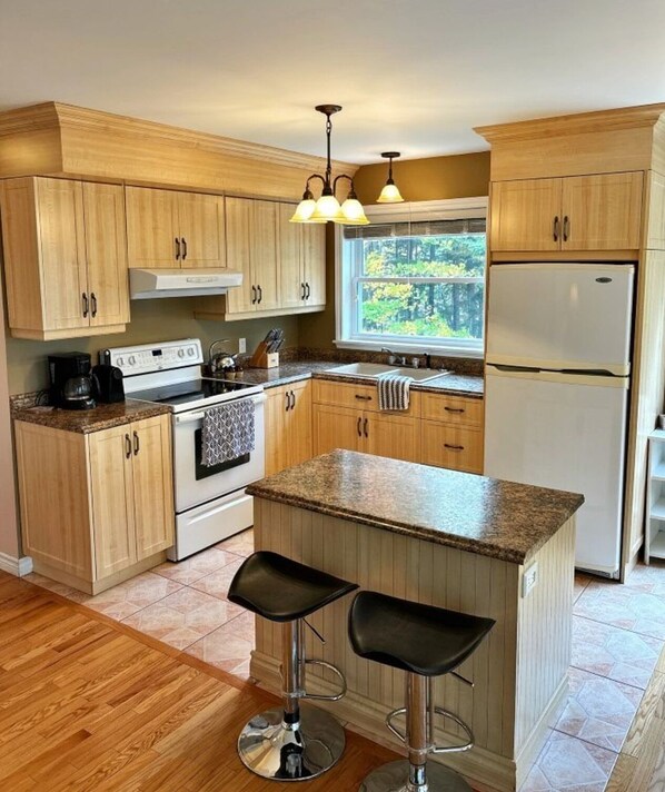 A modern kitchen with wooden cabinets, a central island with bar stools, a refrigerator, and a window with a view of trees. - A Little Cottage by the River in the Heart of the Acadian Peninsula (Pokemouche)