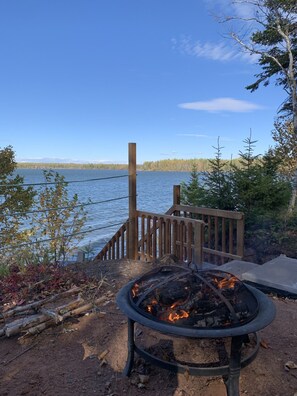 A fire pit with logs, a wooden deck, and a view of a lake and forest. - Riverfront cottage-style home in the heart of the Acadian Peninsula (Pokemouche)