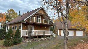 A two-story house with a front porch, a stone foundation, and a garage. - Riverfront cottage-style home in the heart of the Acadian Peninsula (Pokemouche)