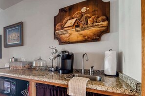 A kitchen with granite countertops, a coffee maker, a sink, and a painting of a rural scene.
