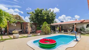 A swimming pool with a red and green inflatable ring, surrounded by potted plants and a house with a tiled roof. - Livia Cristina  (Barra de Santo Antônio)
