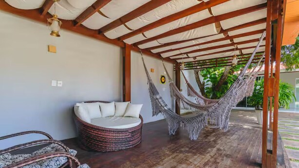 A hammock chair under a wooden pergola, a wicker sofa with cushions, and a wicker chair with a patterned cushion. - Livia Cristina  (Barra de Santo Antônio)