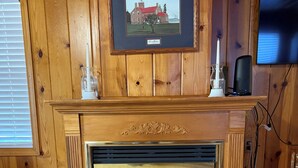Une cheminée surmontée d’un tableau encadré, un téléviseur accroché au mur sur la droite et une chaise en bois placée à droite de la cheminée.