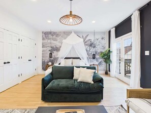 Une pièce avec un canapé vert, un lit à baldaquin, un mur blanc orné d’une fresque et une grande fenêtre.