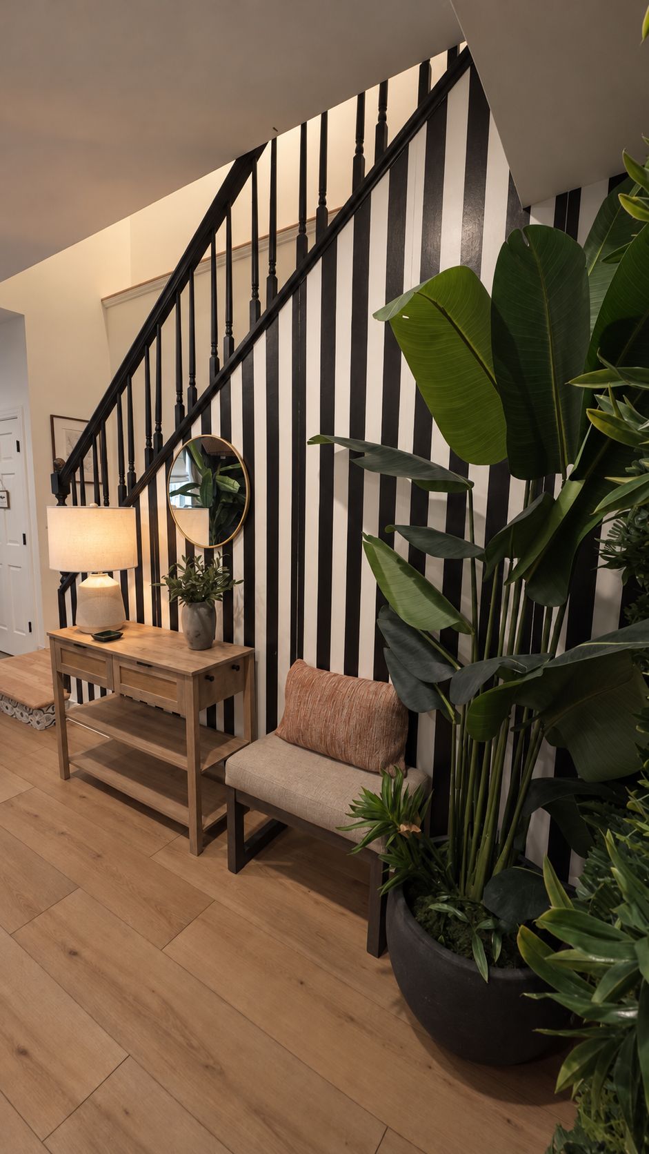 A staircase with black and white striped walls, a wooden bench, a table with a lamp, and large green plants.