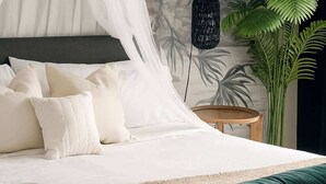 A bed with a canopy, pillows, a woven rug, a green cushion, a wall with a plant mural, and a hanging light fixture.
