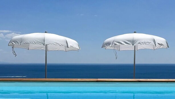Une piscine à l’eau d’un bleu cristallin, deux parasols blancs et une terrasse en bois.