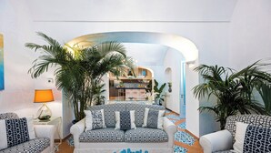 Un salon moderne avec un ensemble de canapés, une table basse et une lampe. On aperçoit des plantes et, au fond, une vue sur la cuisine.