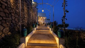 Un sentier bordé de murs en pierre mène à un bâtiment doté d’une entrée couverte, flanqué de grands vases et de plantes.