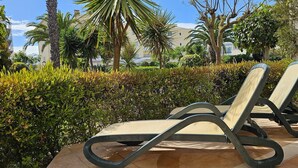 Two lounge chairs with a cushioned seat and backrest, positioned on a tiled patio with a hedge and palm trees in the background. - NEW! Spacious two-bedroom apartment. 2 terraces, pool, 10 minutes to the beach. (Lote 78, Bloco D.)