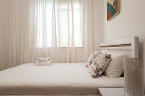 Una cama bien hecha con un almohadón floral, una toalla enrollada y un cabecero con panel de madera.