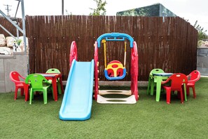 Zona infantil con columpios, tobogán y sillas de plástico.