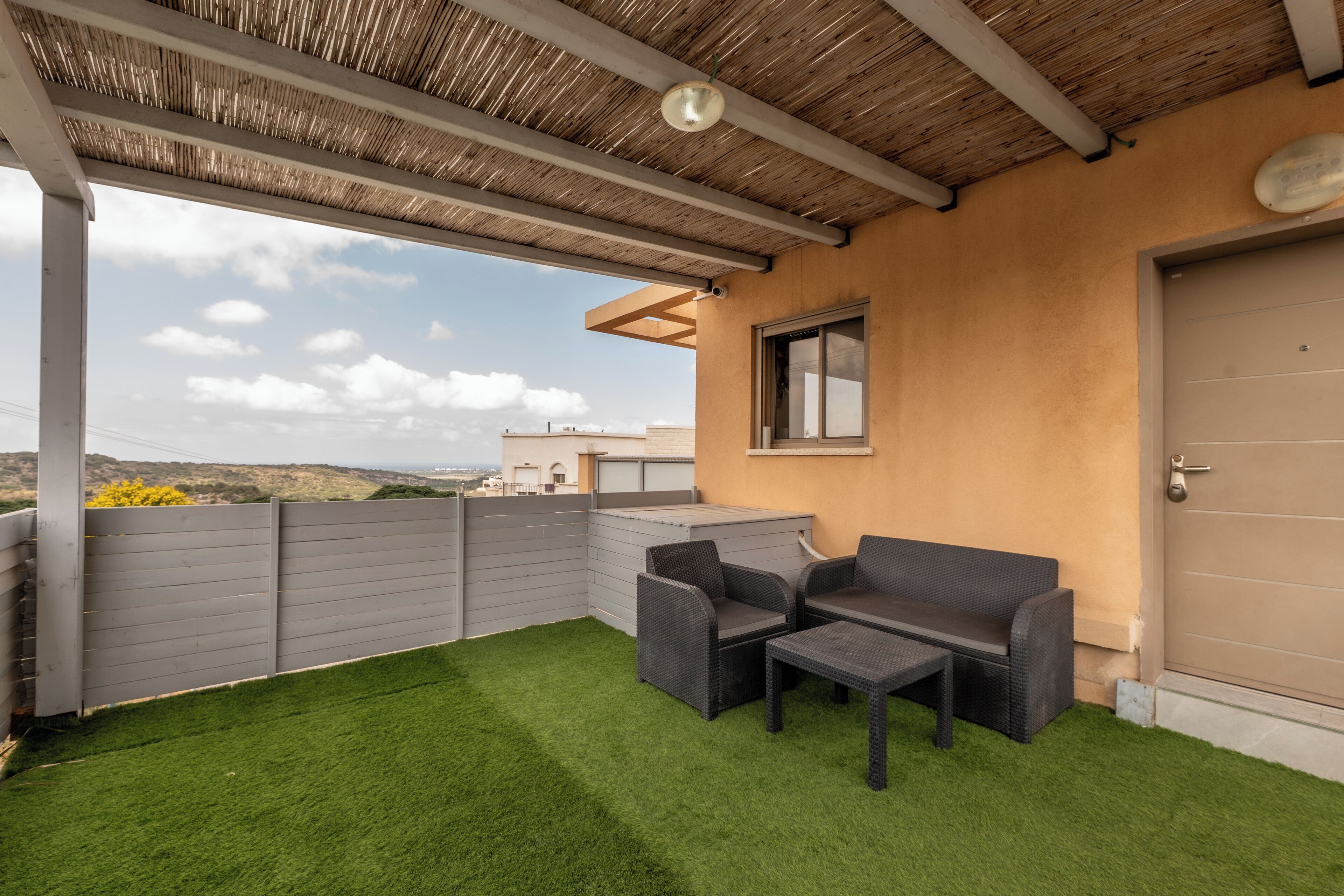 A covered outdoor area with artificial grass, two wicker chairs, a small table, and a door to the right.
