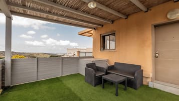 A covered outdoor area with artificial grass, two wicker chairs, a small table, and a door to the right.