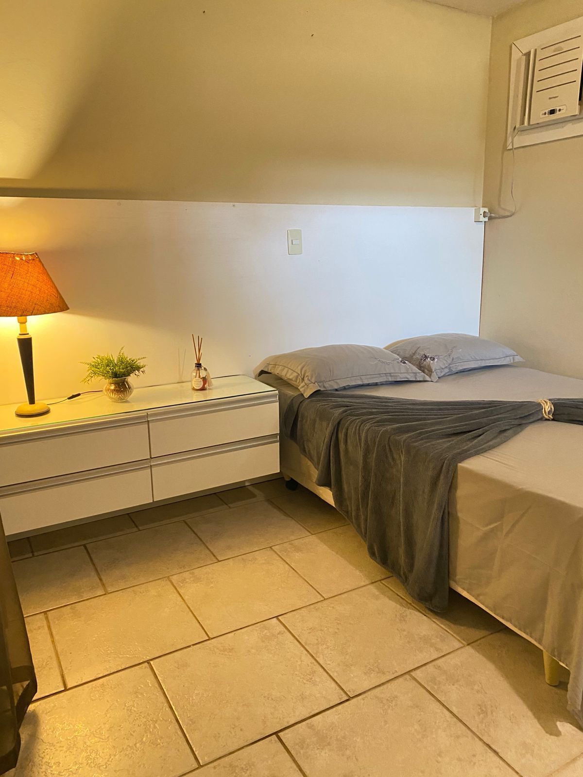 Une petite chambre bien éclairée, avec un lit simple, une table de chevet, une lampe et un climatiseur fixé au mur.