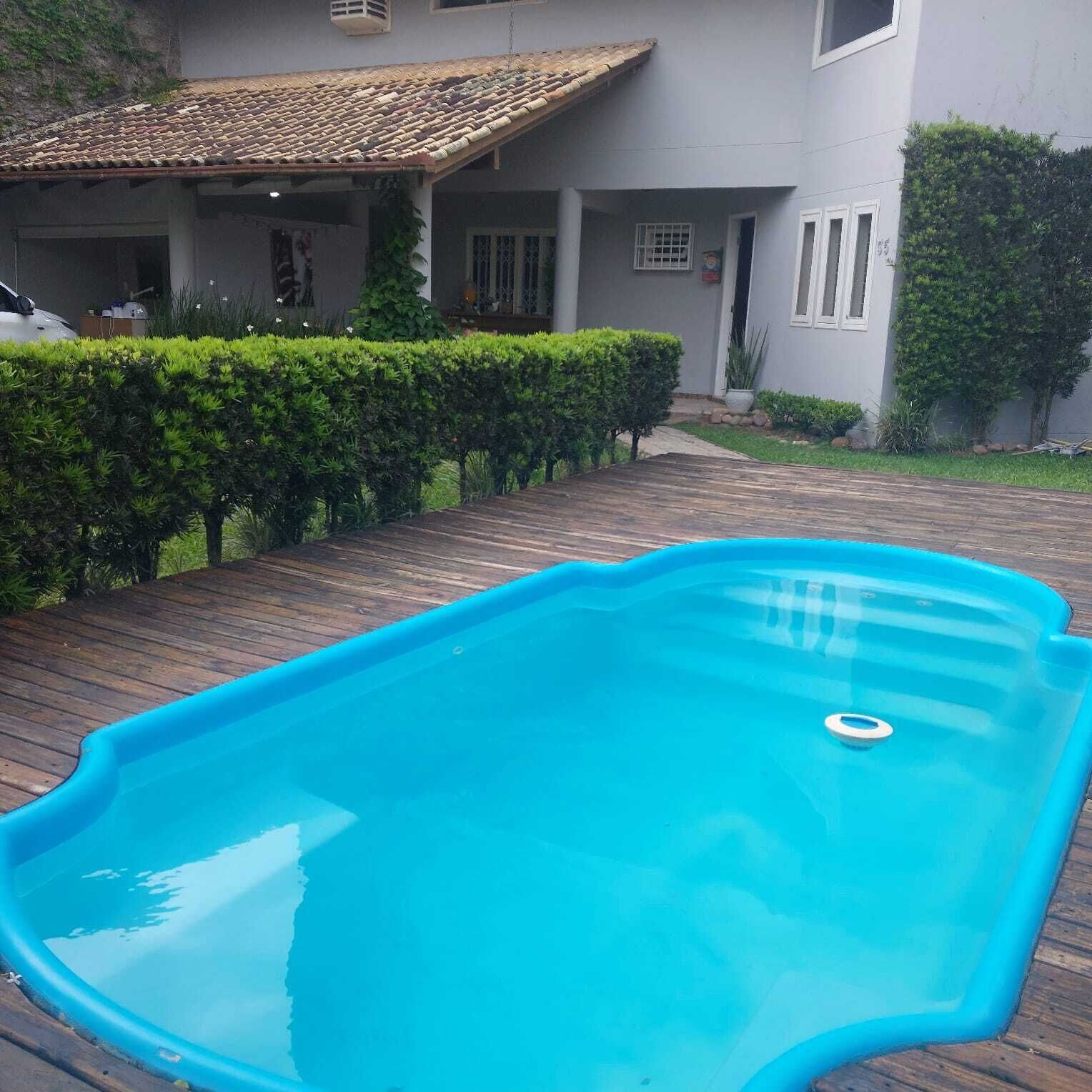 Piscina com deck de madeira, rodeada por um hedge bem cuidado e uma casa com telhado em telhas.