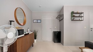 Un salon moderne avec un meuble en bois, un ventilateur blanc, une poubelle noire, un canapé gris et une petite table en bois.