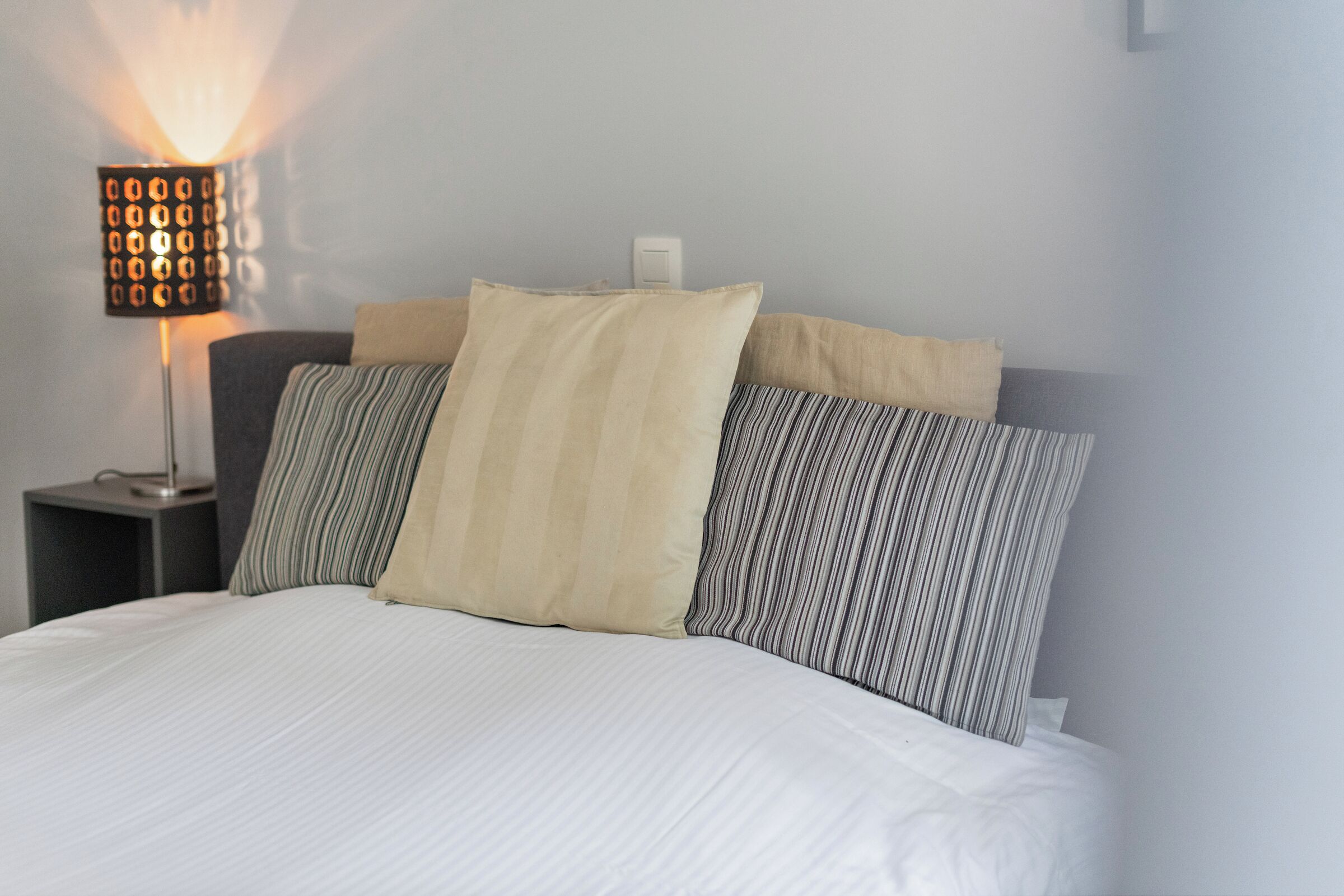 Een net opgemaakt bed met witte beddengoed en gestreepte kussens, een moderne lamp op een nachtkastje, en een effen muur op de achtergrond.