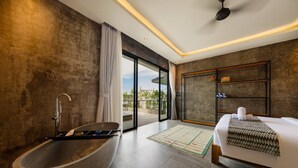 A modern hotel room with a large stone bathtub, a bed with a patterned throw, a wooden headboard, and a view of the outdoors through a sliding glass door.